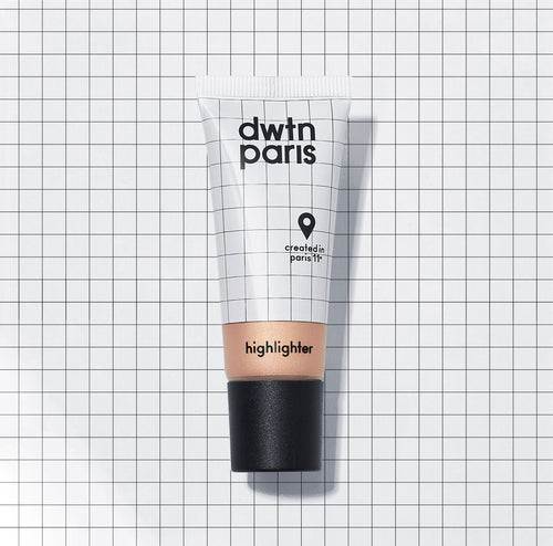Photo du tube de la teinte light du highlighter. Le fond de la photo est blanc avec un quadrillage fin noir. Le fond s'accorde avec le packaging qui lui aussi contient un quadrillage. Une partie transparente laisse voir la couleur beige nacrée du highlighter