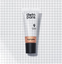 Photo du tube de la teinte medium du highlighter. Le fond de la photo est blanc avec un quadrillage fin noir. Le fond s'accorde avec le packaging qui lui aussi contient un quadrillage. Une partie transparente laisse voir la couleur légèrement cuivrée nacrée du highlighter